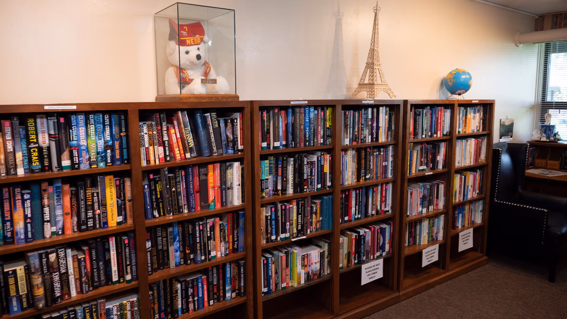Wooden bookshelves filled with books along a wall in a cozy reading area, with a globe, a teddy bear in a display case, and a leather armchair.