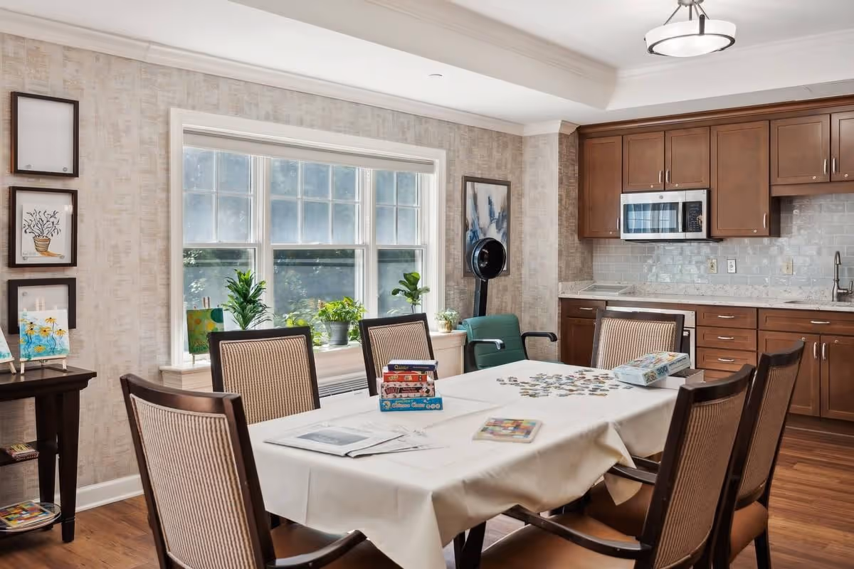 A bright and cozy dining area with a rectangular table covered with a white tablecloth surrounded by six chairs. On the table are board games, a puzzle, and some books. The room has a large window with several potted plants on the windowsill, light-colored wallpaper, and wooden flooring. To the right is a kitchen area with wooden cabinets, a microwave, and a sink. There are framed artworks on the walls and a standing fan in the corner.