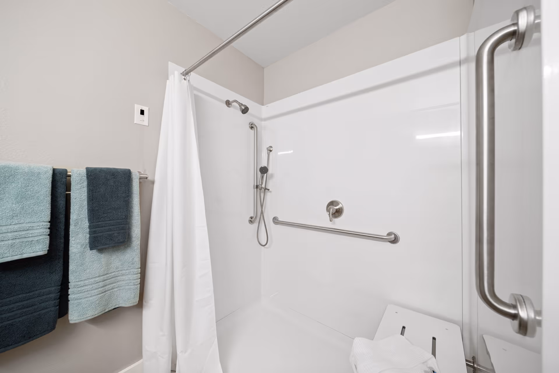 A clean, accessible shower area with white walls and a white shower curtain. The shower features stainless steel grab bars, a handheld showerhead, and a fold-down shower seat with a white towel on it. On the left wall, there are three towels hanging on a towel rack in shades of blue and teal.