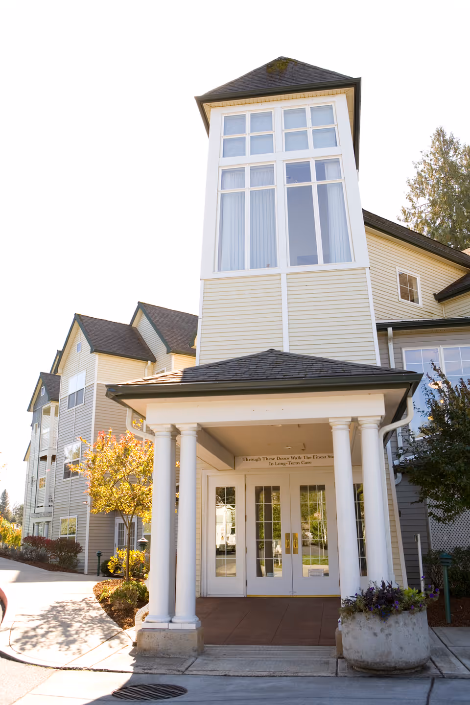 Entrance of a multi-story senior living facility with a covered porch supported by four white columns. The building has beige siding and large windows, with a small tree and plants near the entrance.