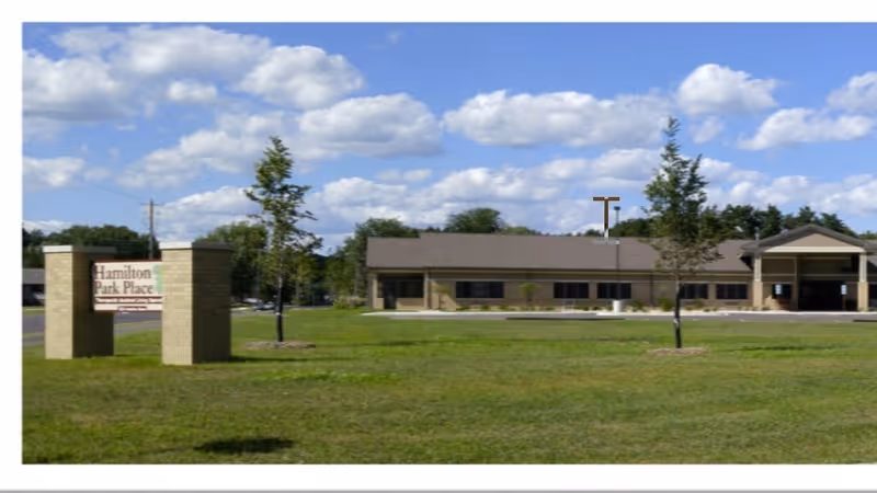 Exterior view of Hamilton Park Place Assisted Living facility with a large grassy lawn, a few young trees, and a clear blue sky with scattered clouds. The building is single-story with a brown roof and beige walls. A sign with the facility name is visible on the left side of the image.