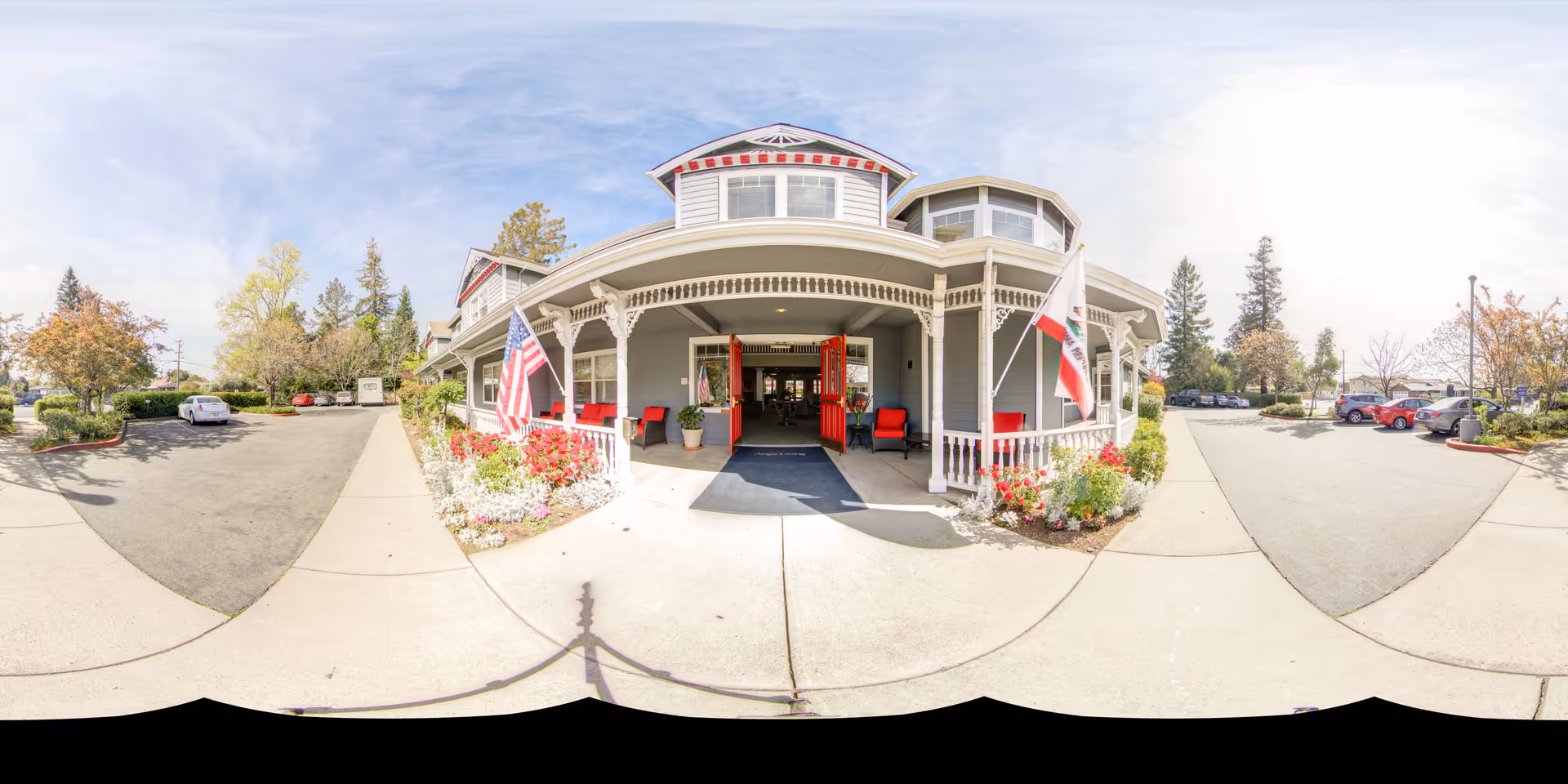 Front exterior view of Aegis Living Napa facility with a covered porch entrance, American and California state flags, red chairs, flower beds, and a parking lot with several cars under a partly cloudy sky.