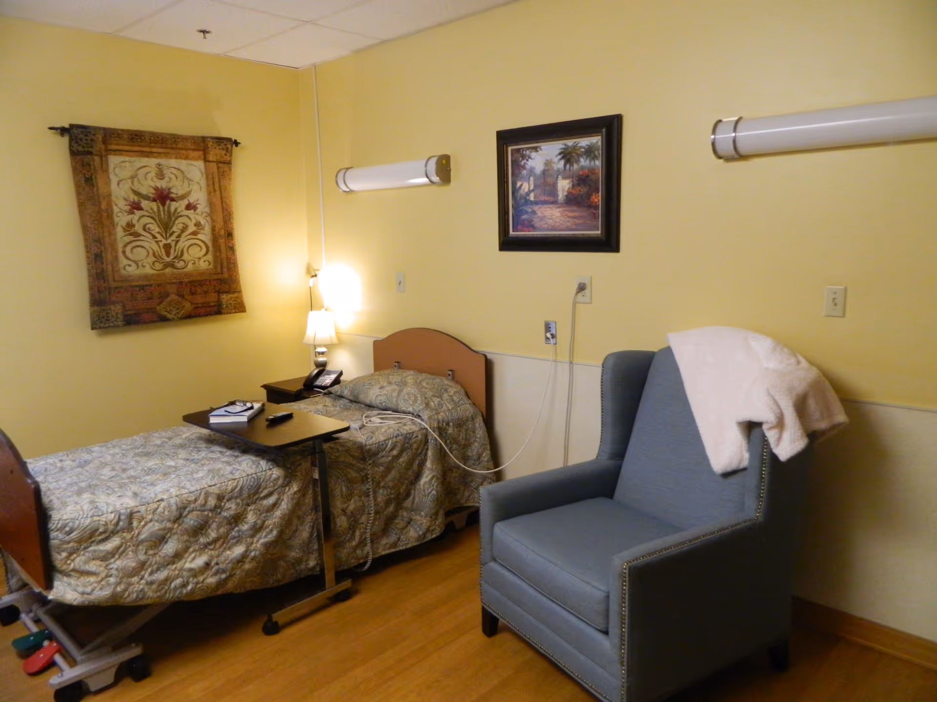 A nursing facility bedroom with a single hospital-style bed covered in a patterned bedspread, a small overbed table with a remote and book, a bedside table with a lamp and phone, a blue upholstered armchair with a white blanket draped over the back, and two framed wall hangings on yellow walls.
