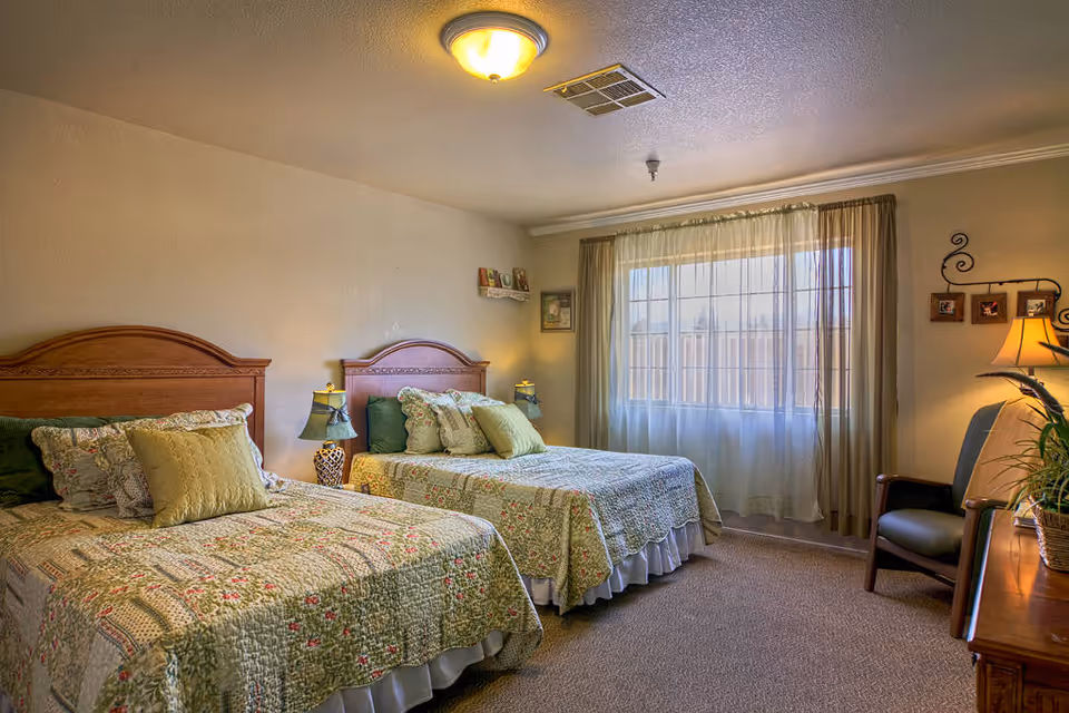 A cozy bedroom with two twin beds featuring wooden headboards and floral patterned bedspreads. Each bed has multiple pillows in green and beige tones. Between the beds is a small nightstand with a decorative lamp. A large window with sheer curtains allows natural light to fill the room. On the right side, there is a wooden dresser with a potted plant and a chair next to it. The walls are decorated with framed pictures and a small shelf holding books.