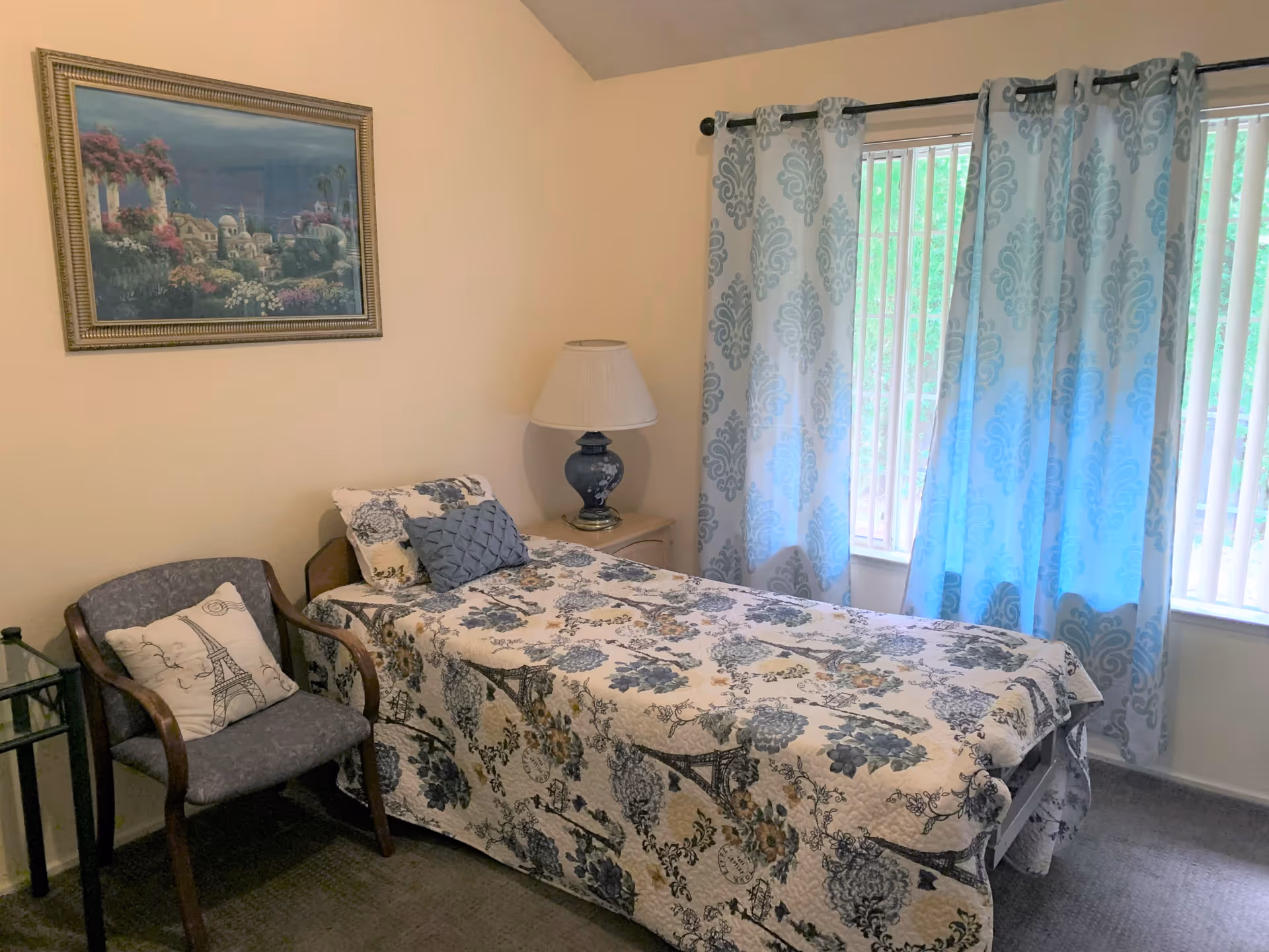 Small furnished bedroom with a single bed covered in a patterned quilt, a bedside lamp, chair, and window with patterned curtains.