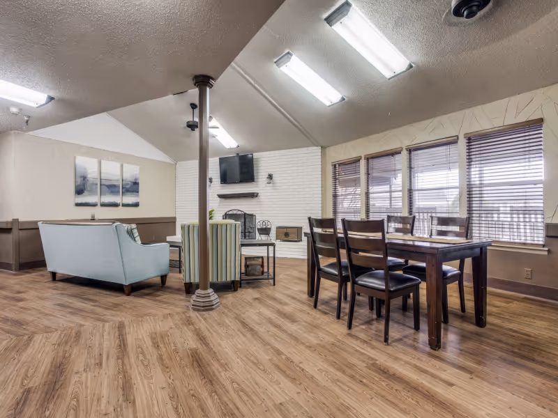 A spacious senior living common area featuring a wooden dining table with six chairs on the right side, and a seating area with a light blue sofa and a striped armchair on the left. The room has wood flooring, a white brick fireplace with a mounted TV above it, and several windows with blinds letting in natural light.