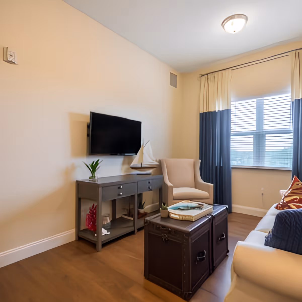 A cozy living room with a mounted flat-screen TV above a gray console table, a beige armchair, a window with blue and beige curtains, and a white sofa with colorful pillows. A dark wooden trunk serves as a coffee table with books and small plants on top.