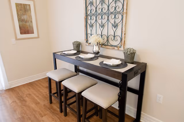 Narrow console-style dining table with three cushioned stools, place settings, and decorative wall art above.