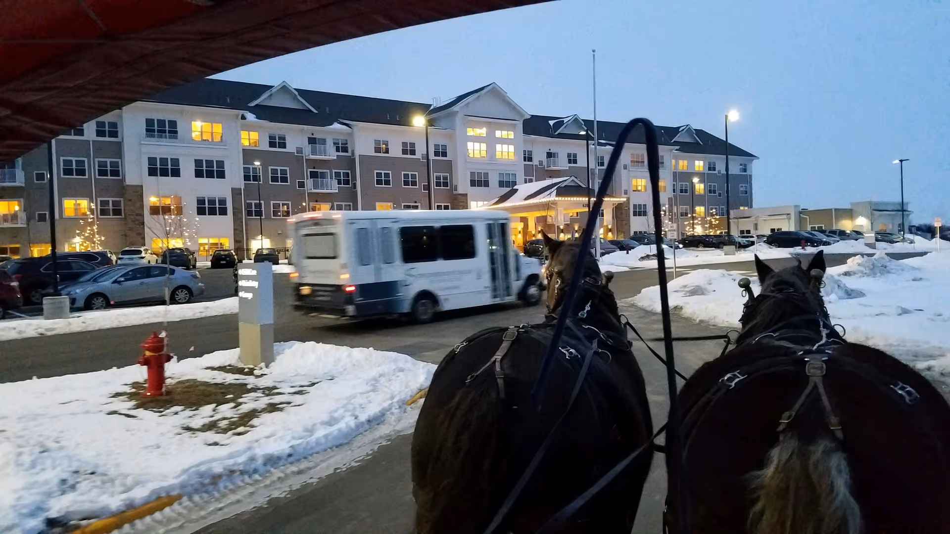 View of Riley Crossing Senior Living building exterior at dusk with snow on the ground, a shuttle bus driving by, and two horses harnessed in the foreground.