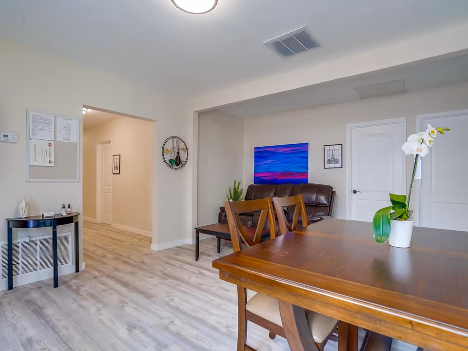 Interior view of a senior living facility room featuring a wooden dining table with chairs, a white orchid plant on the table, a brown leather couch against the wall, colorful artwork above the couch, light wood flooring, and a small black side table with decorative items near a hallway entrance.