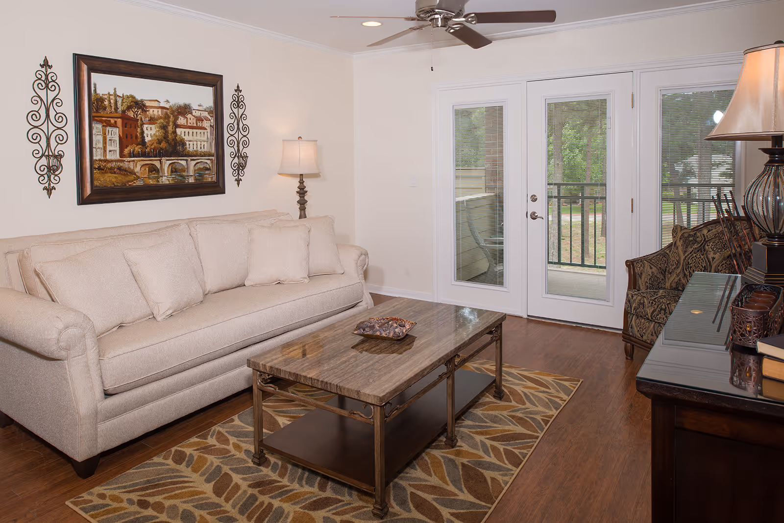 A cozy living room with a beige sofa, a patterned armchair, a wooden coffee table on a leaf-patterned rug, a floor lamp, and a ceiling fan. There is a framed painting and decorative wall sconces above the sofa. French doors with glass panels lead to an outdoor balcony with greenery visible outside.