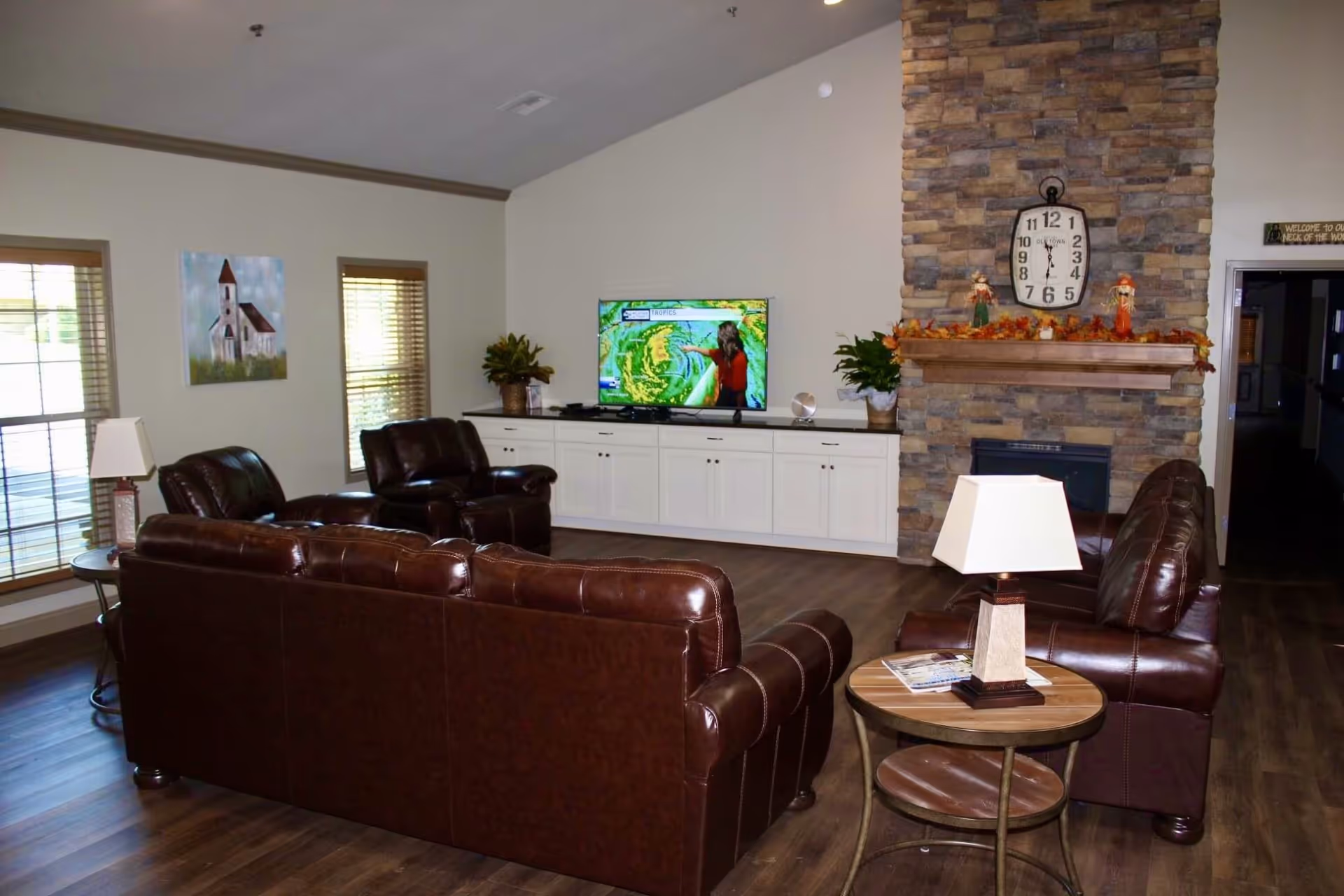 A furnished living room with brown leather sofas and chairs facing a TV on white cabinets and a stone fireplace with a mantel clock.