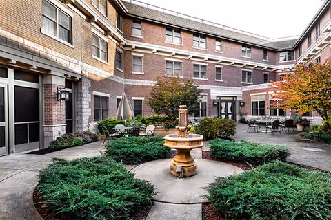 Outdoor courtyard area of a senior living facility with a central stone fountain surrounded by green shrubs and paved walkways. The courtyard is enclosed by a three-story brick building with multiple windows and outdoor seating areas with tables and chairs. Trees with autumn-colored leaves are visible on the right side.