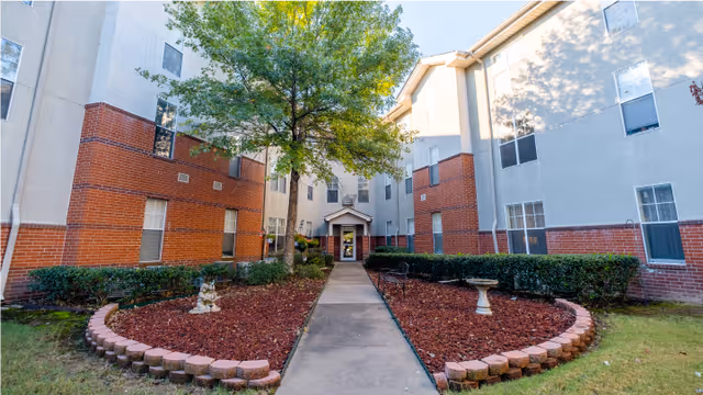 Exterior view of a multi-story retirement facility building with a central walkway leading to the entrance. The building has a combination of beige and red brick walls, several windows, and a tree with green leaves in the landscaped area along the walkway.