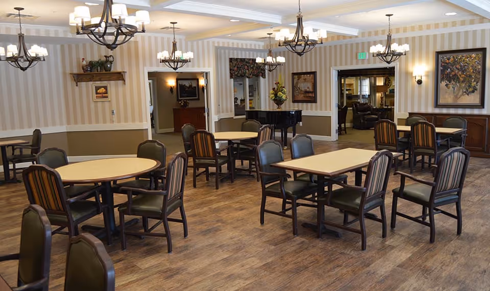 Spacious dining room with round and rectangular tables, chairs, chandeliers, a piano and framed artwork on the walls.