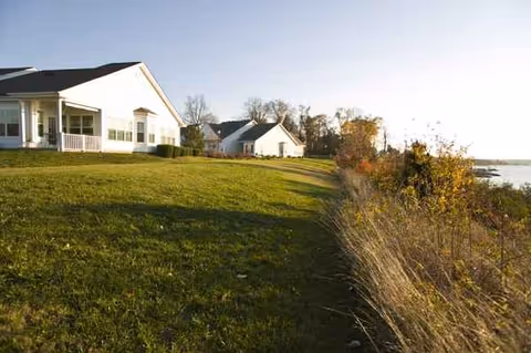 White senior living buildings line a grassy waterfront lawn beside a lake.