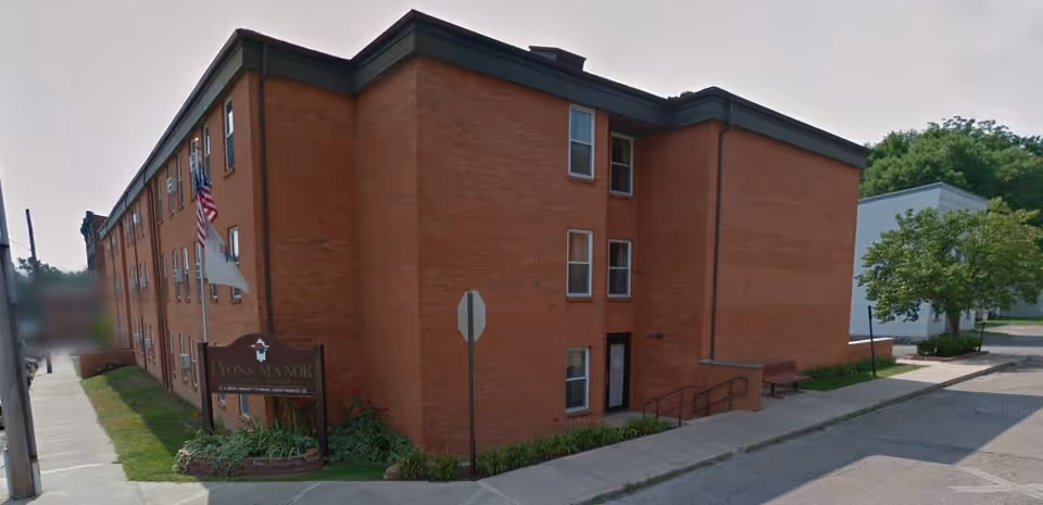 Exterior view of a three-story brick building with multiple windows, a sidewalk, and a sign that reads Lyons Manor. There are two flags on poles near the sign, and some greenery around the building. The street and a tree are visible on the right side.