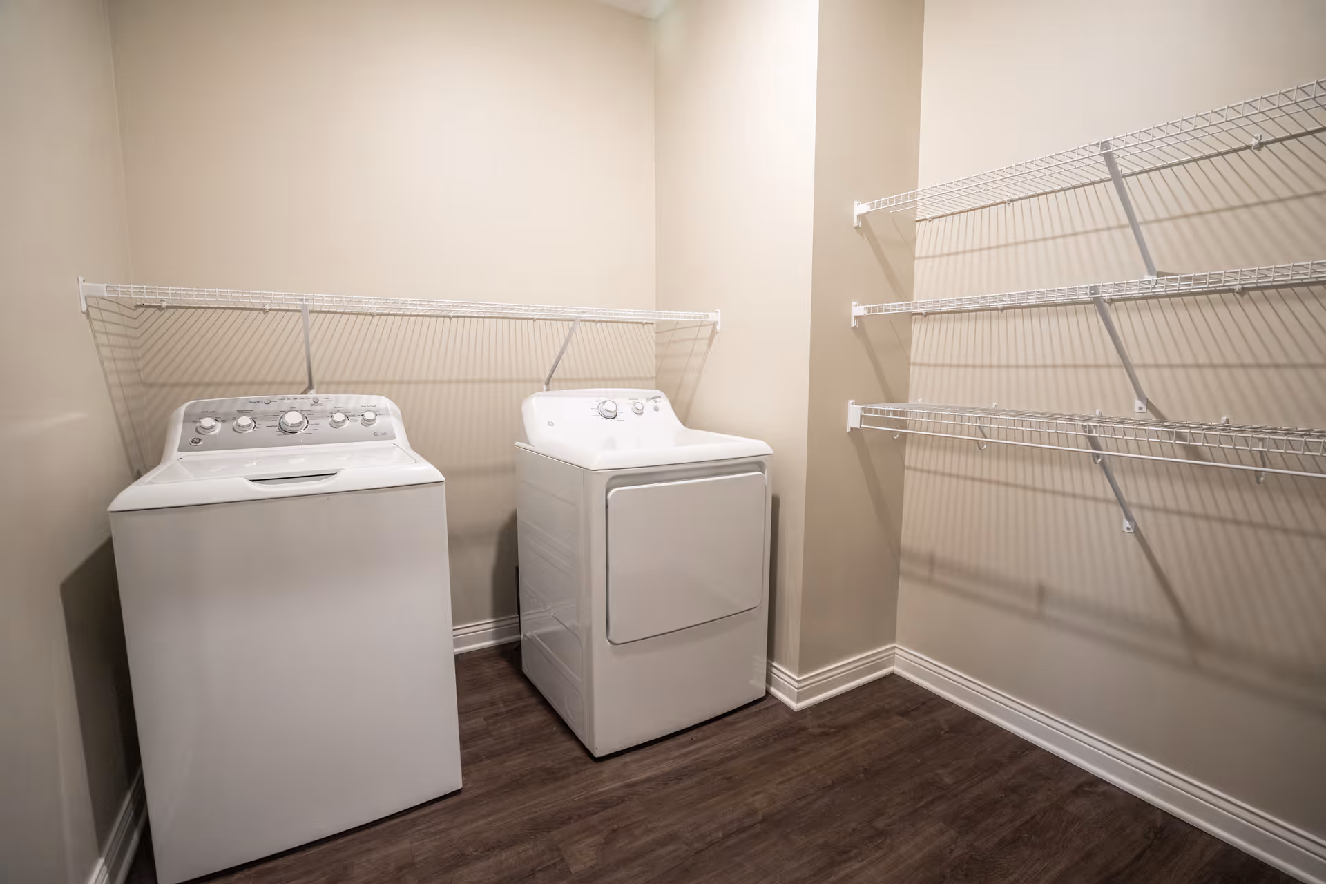 Laundry room with a white top-loading washing machine and matching dryer beneath wire shelving and laminate floors.