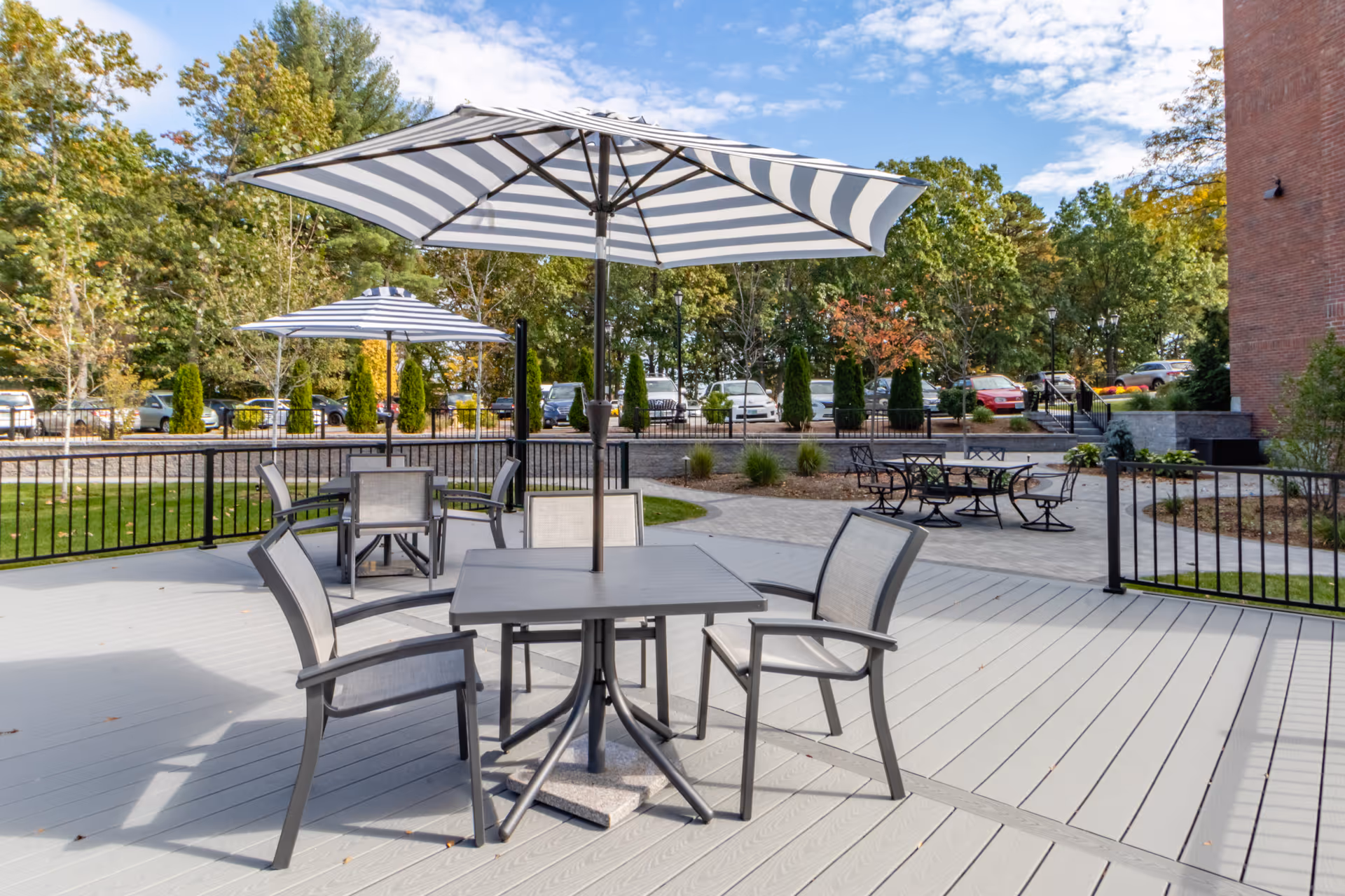 Outdoor patio area with multiple tables and chairs under large striped umbrellas, surrounded by trees and a parking lot in the background.