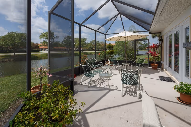 Screened lakefront patio with outdoor chairs, tables, an umbrella, and potted plants.
