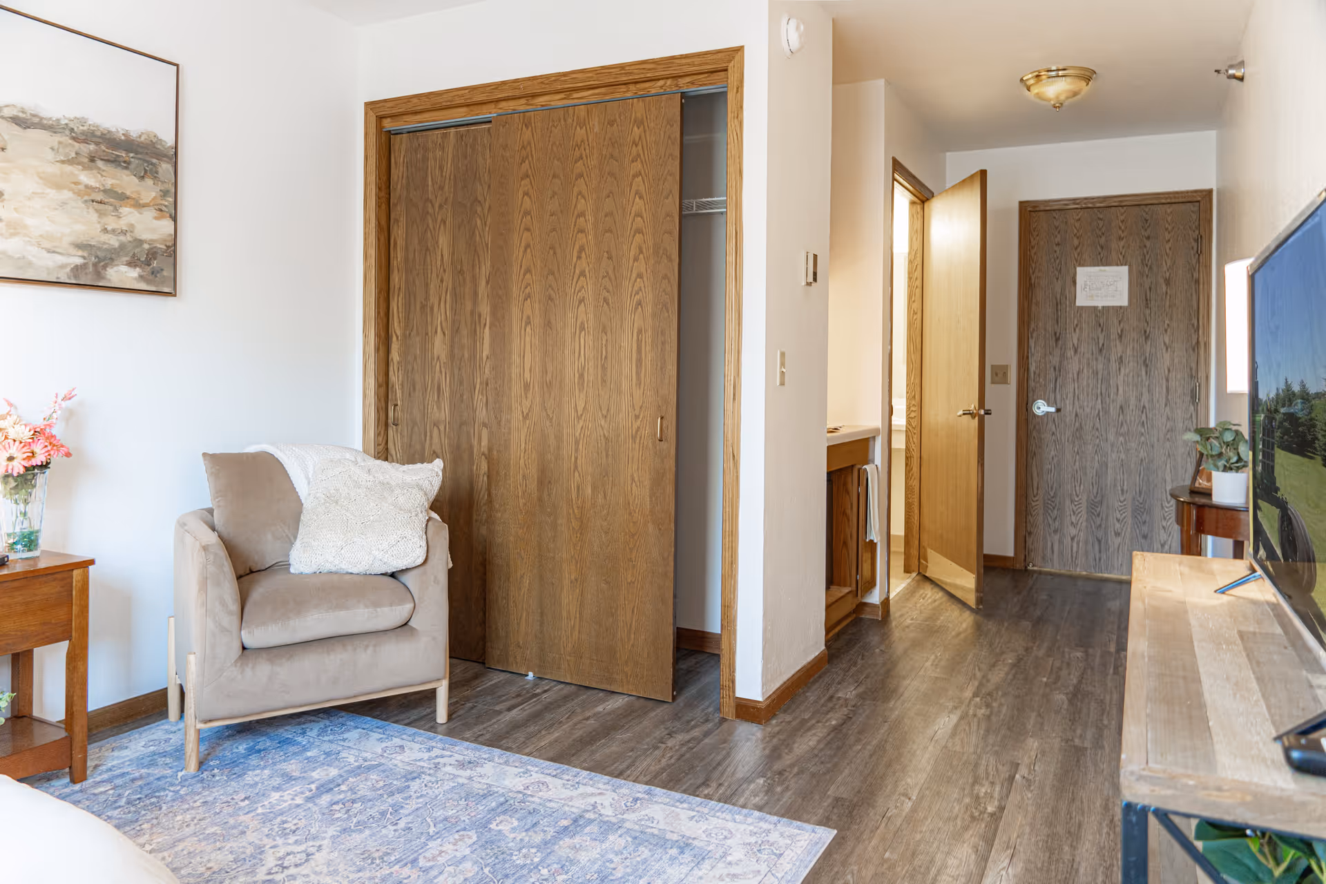 A cozy living area with a beige armchair holding a white knitted pillow, a wooden side table with a vase of pink flowers, a large wooden closet with sliding doors, and a TV on a wooden stand. The room has wood flooring, white walls, and a hallway leading to a door and a bathroom vanity area.