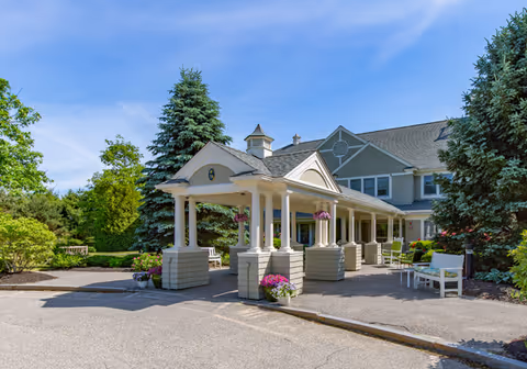 Covered porte-cochère and front entrance of a senior living building with benches, potted flowers, and surrounding trees.