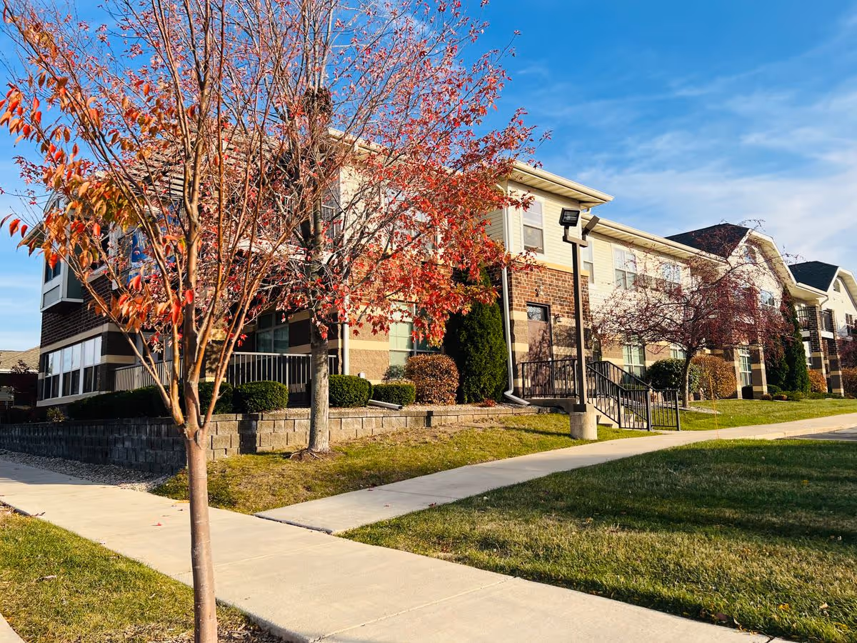 Two-story senior living building exterior with red-leaved trees and a sidewalk in front.