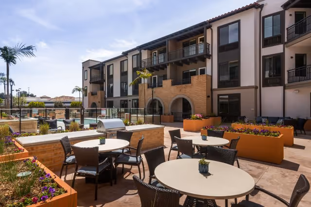 Outdoor courtyard patio with round tables, chairs, planters and a grill in front of a three-story senior living building with balconies.