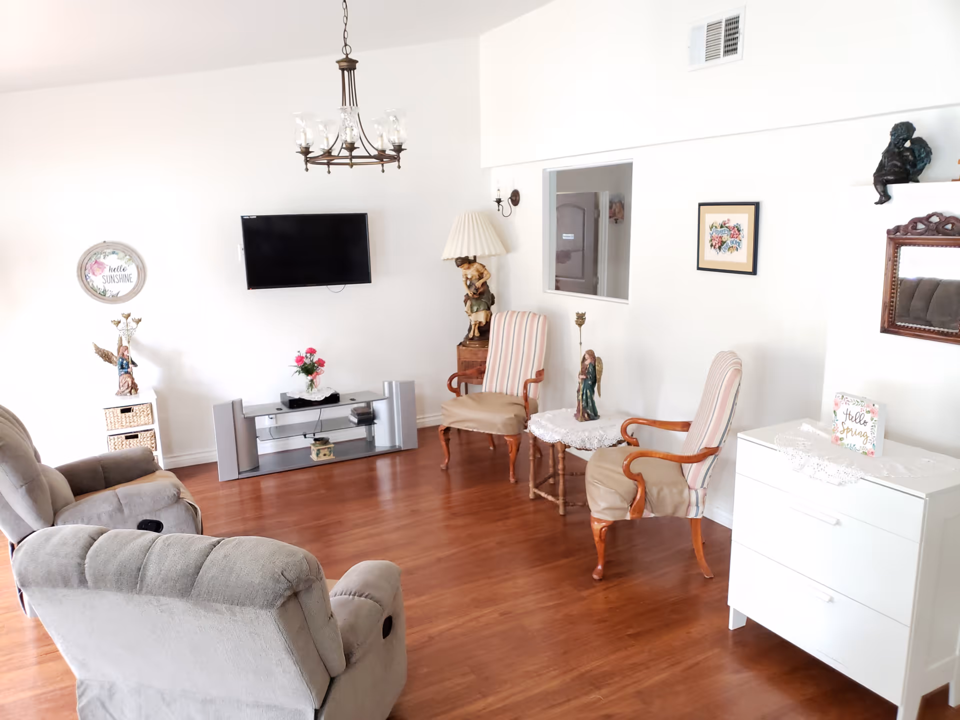 A bright living room with wooden flooring featuring two beige recliner chairs, two striped armchairs, a small round table with a lace cover and a figurine, a white dresser with a decorative card, a wall-mounted flat-screen TV above a modern TV stand with speakers and a flower vase, wall decorations including framed art and a mirror, and a chandelier hanging from the ceiling.