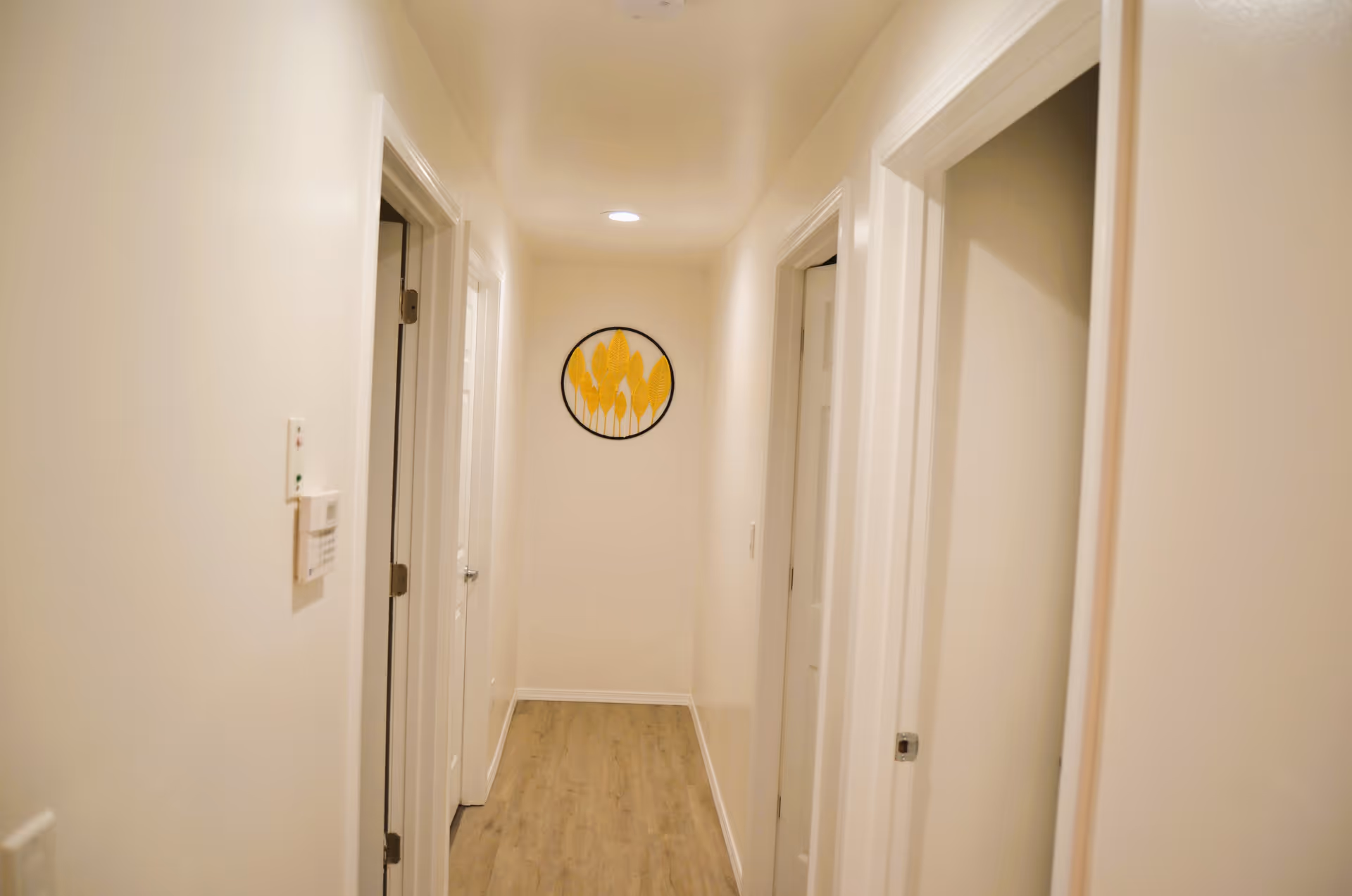 A narrow hallway with light-colored walls and wooden flooring. There are three white doors visible, two on the right and one on the left. A round wall decoration with yellow leaf patterns is hung at the end of the hallway. The ceiling has recessed lighting.