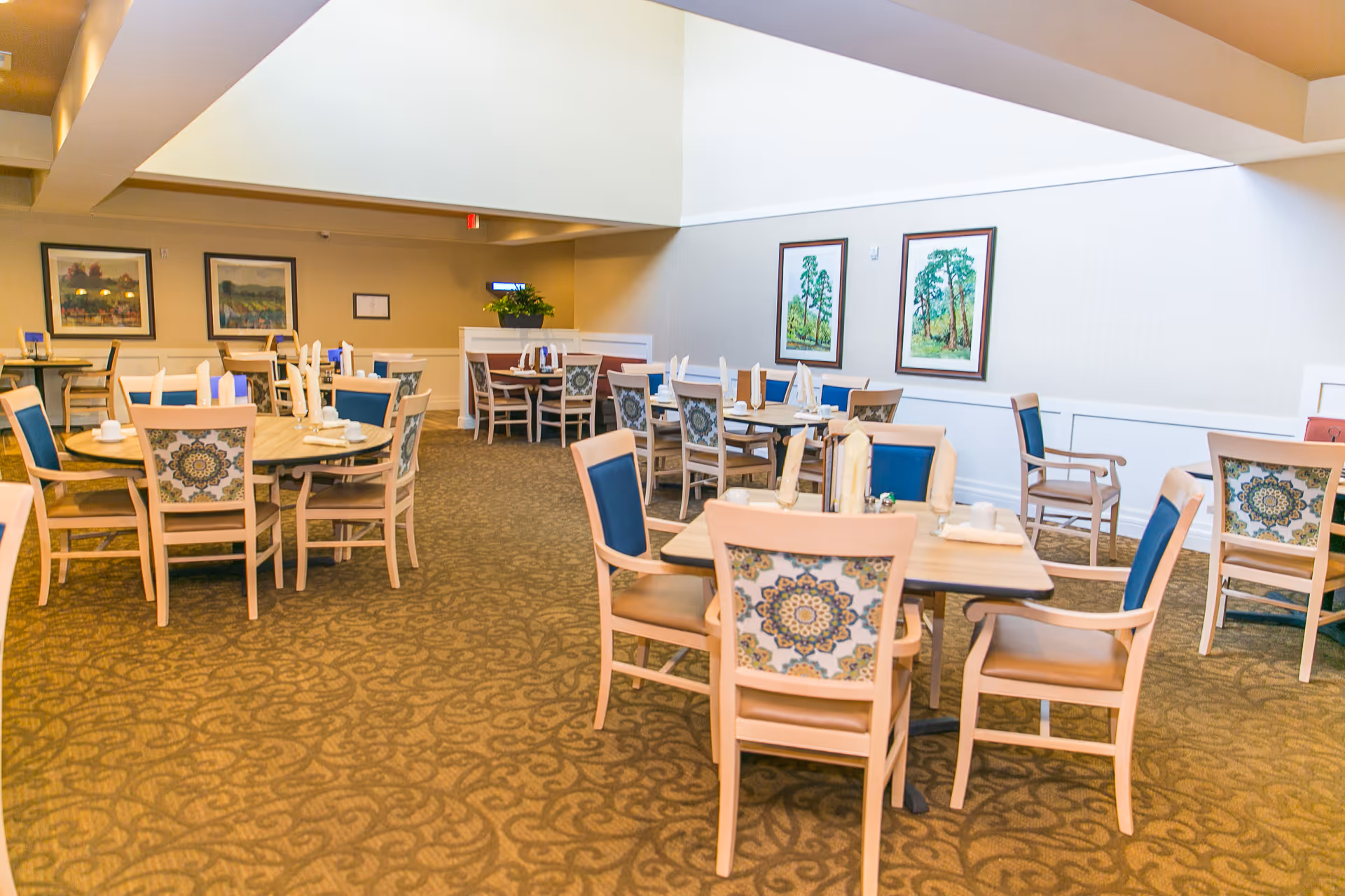 A spacious dining room in a senior living facility with multiple tables and chairs arranged neatly. The chairs have patterned backs and blue cushions. The room is well-lit with natural light coming from a skylight above. There are framed paintings of trees on the walls and a carpeted floor with a decorative pattern.