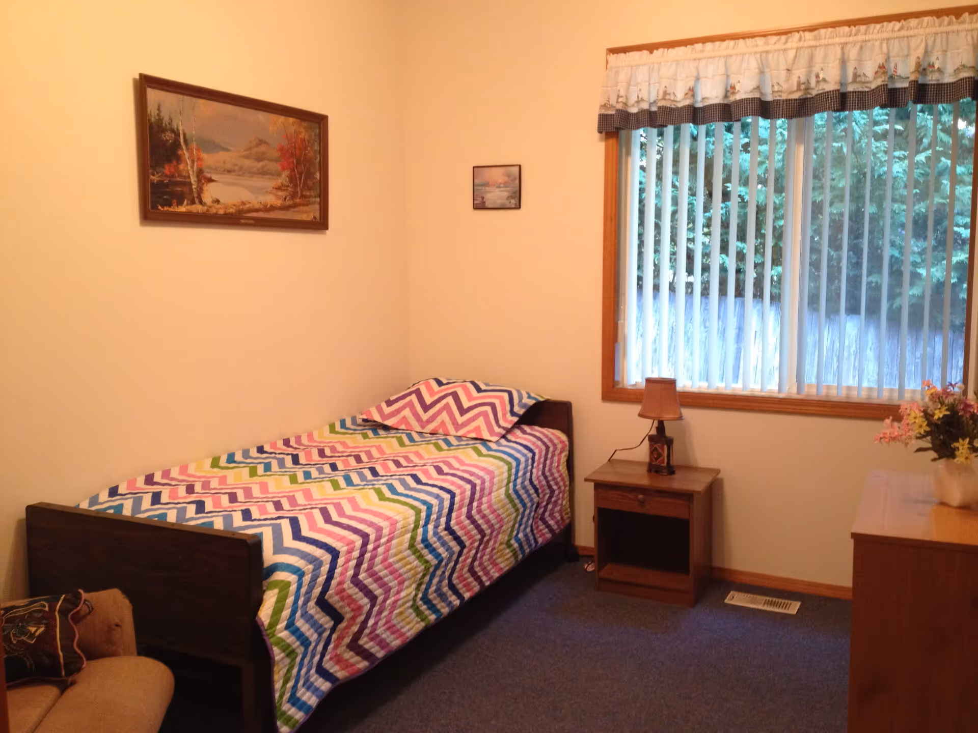 A small bedroom with a single bed covered in a colorful zigzag patterned quilt. There is a wooden nightstand with a lamp next to the bed, a window with vertical blinds and a valance, a wooden dresser with a vase of flowers, and two framed landscape paintings on the walls.