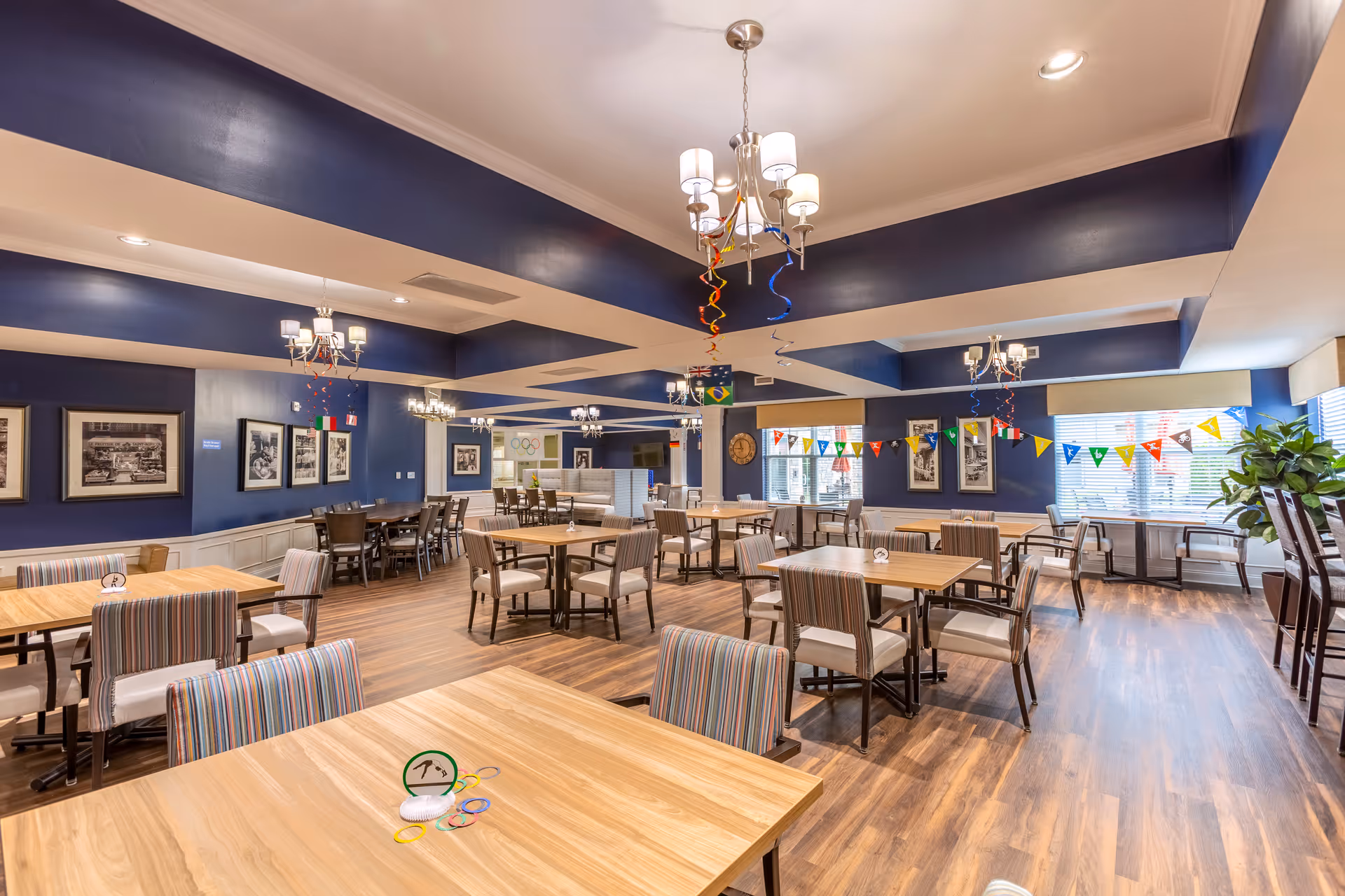 A spacious dining room with wooden floors and multiple tables and chairs arranged neatly. The walls are painted dark blue with white wainscoting, and decorated with framed black and white photographs. Colorful pennant banners and hanging spiral decorations add a festive touch. Large windows allow natural light to fill the room.