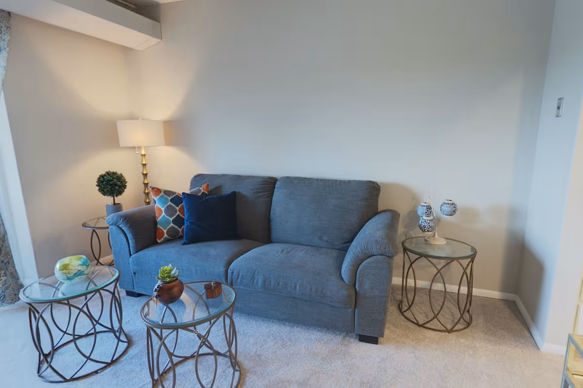 A cozy living room area with a gray upholstered sofa adorned with three decorative pillows in blue and patterned colors. There are three round glass-top side tables with metal bases, one holding a small potted plant and another with decorative items. A floor lamp with a white shade and a small artificial topiary plant are placed beside the sofa. The room has light-colored carpet and plain light walls.