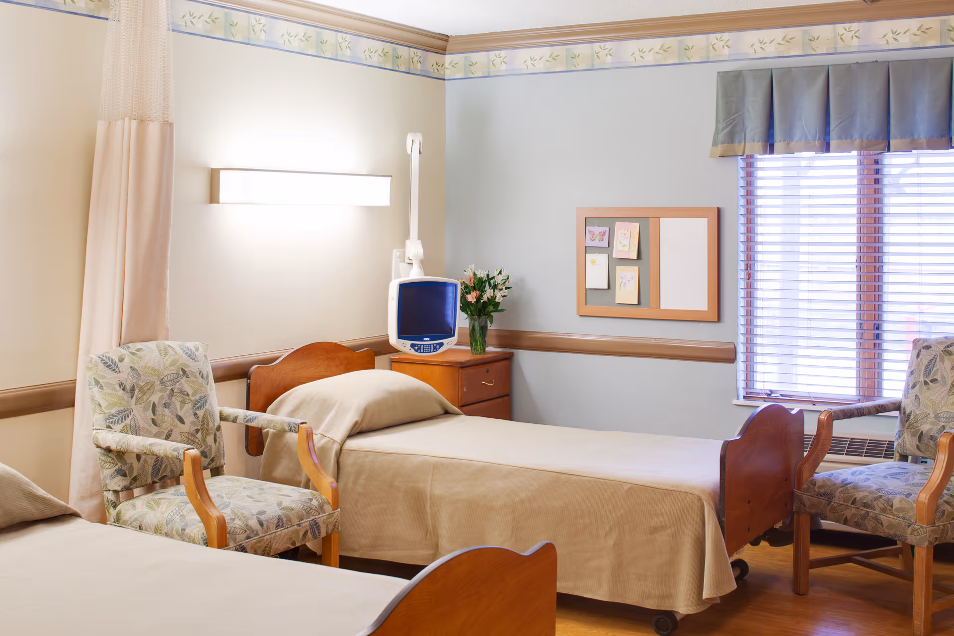 A well-lit room with two single beds covered in beige bedspreads, two floral-patterned armchairs, a wooden nightstand with a small monitor and a vase of flowers, a bulletin board with notes on the wall, and a window with blinds and a blue valance.