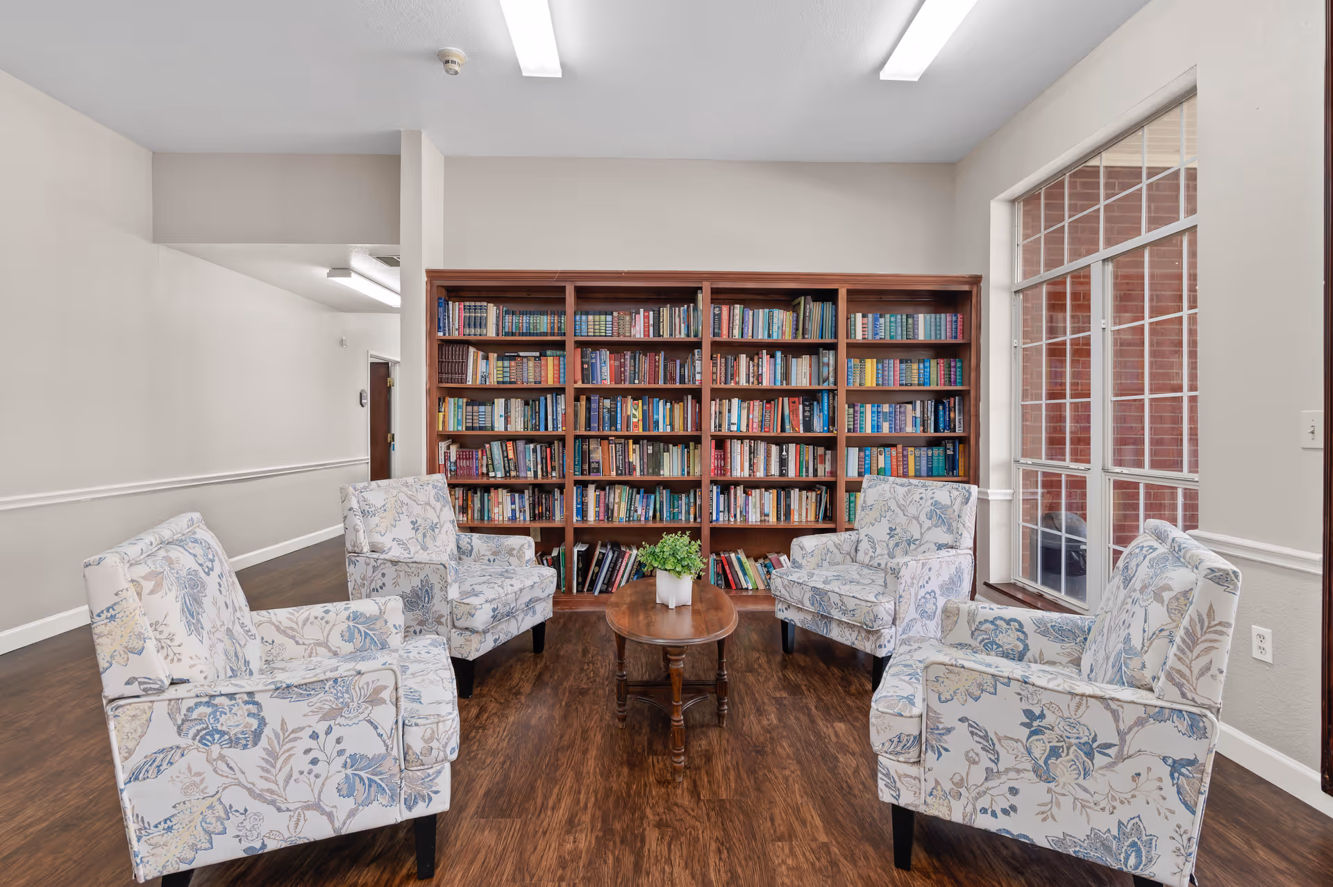 A cozy reading area in an assisted living facility featuring four floral-patterned armchairs arranged around a small wooden coffee table with a potted plant. Behind the seating is a large wooden bookshelf filled with books. To the right, there is a large window with a view of a brick exterior wall.