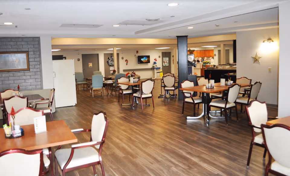 A spacious dining room with multiple wooden tables and cushioned chairs arranged neatly. The room has wood flooring, soft lighting, and a visible kitchen area in the background. There are decorations on the walls including a star and a framed picture.