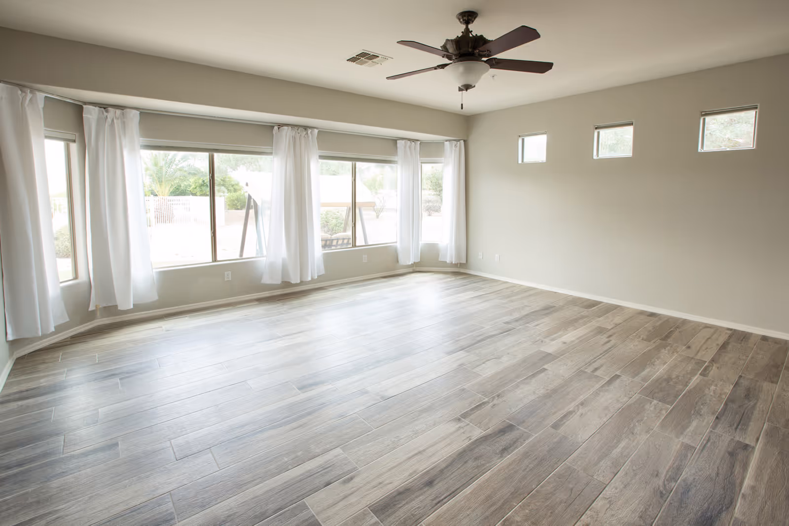 Empty room with light wood-look tile flooring, beige walls, a ceiling fan with light, and large windows with white curtains letting in natural light. Small square windows are near the ceiling on one wall.