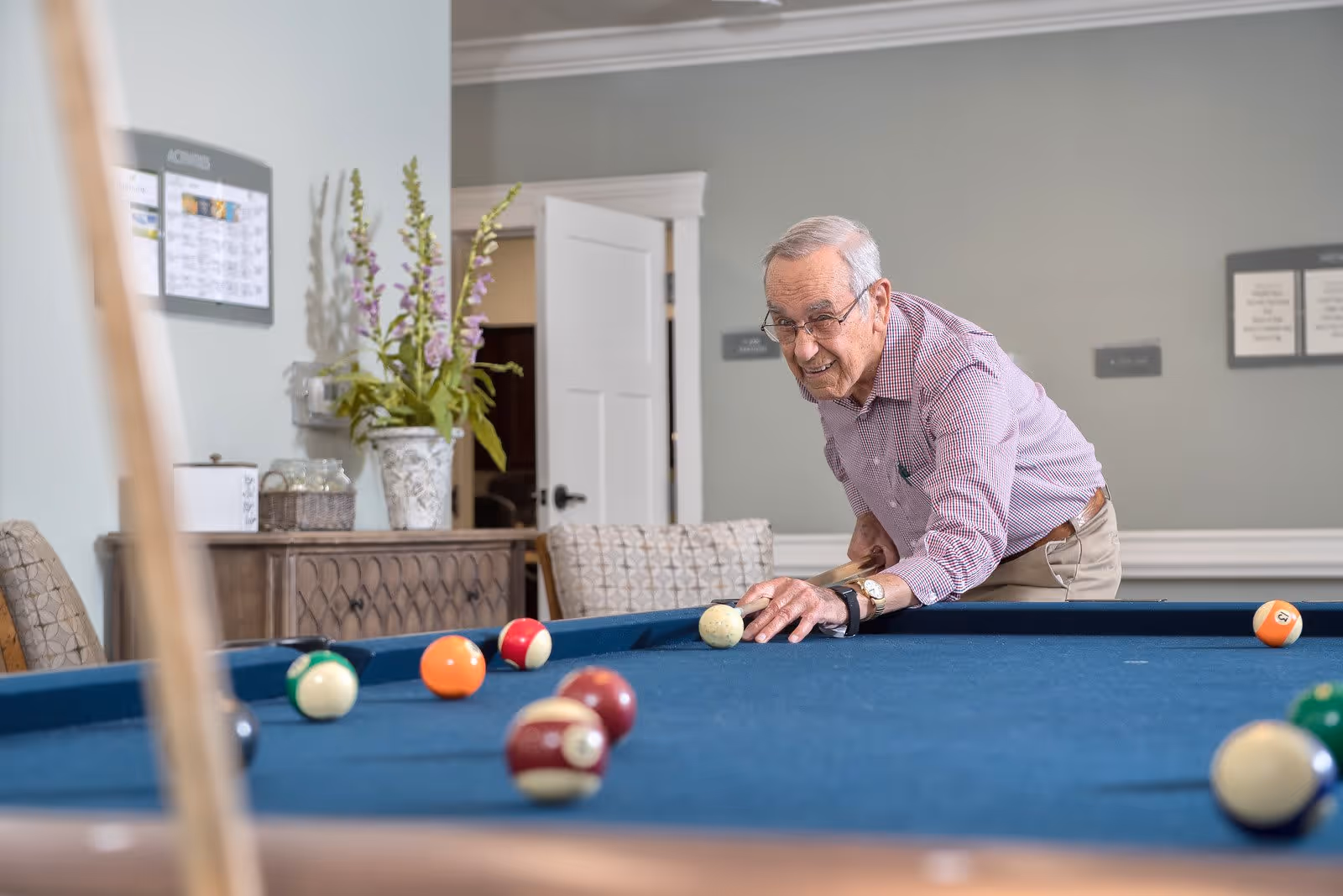 An elderly man wearing glasses and a checkered shirt is playing pool on a blue felt pool table in a well-lit room with light gray walls. Behind him, there is a wooden cabinet with a vase of flowers and a wall calendar labeled 'ACTIVITIES'. The room has a cozy and inviting atmosphere.