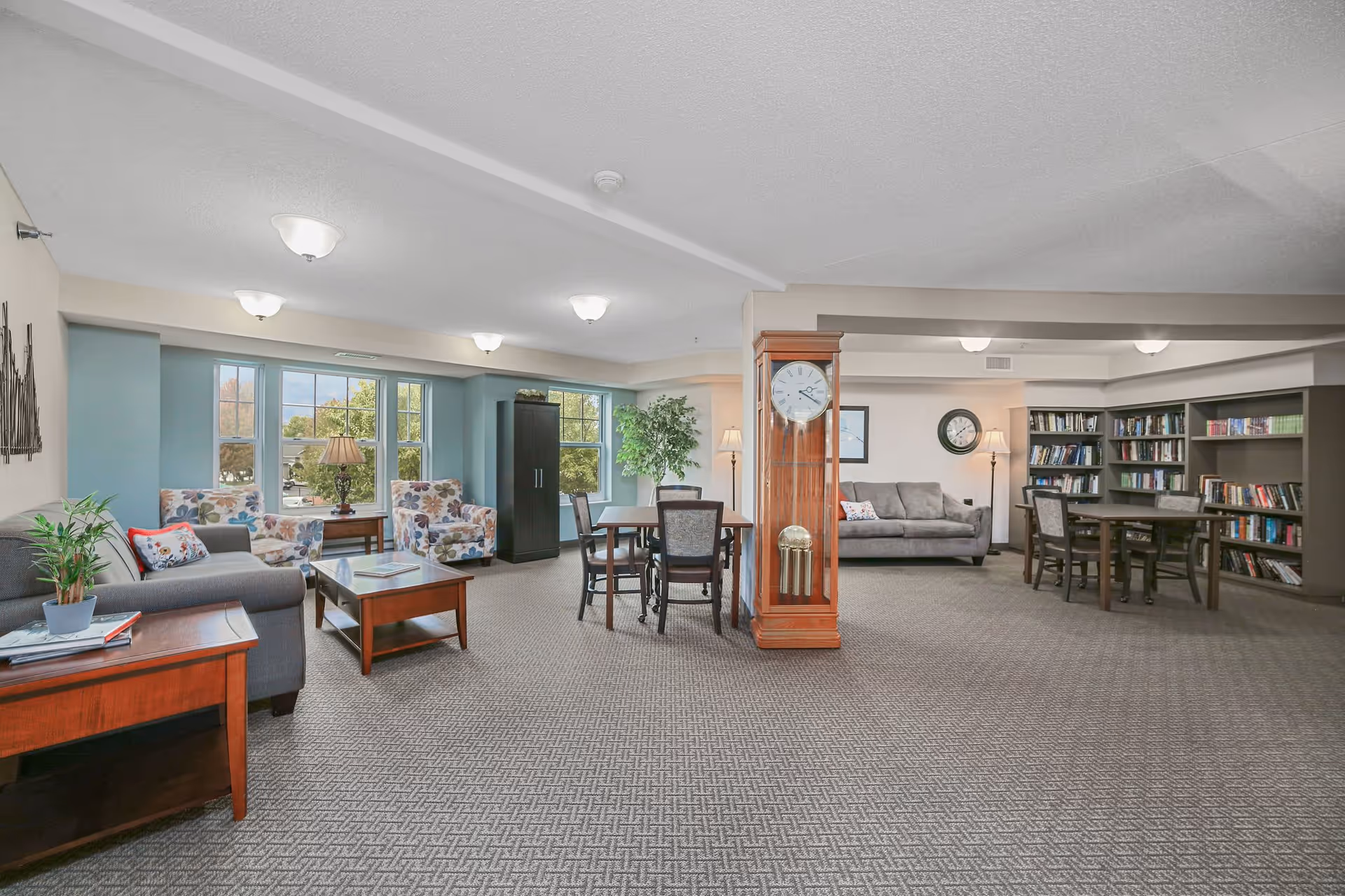 A spacious senior living facility common area with multiple seating arrangements including sofas, floral armchairs, and tables with chairs. There is a large wooden grandfather clock in the center, bookshelves filled with books along the back wall, and large windows letting in natural light. The room has carpeted flooring and soft ceiling lights.