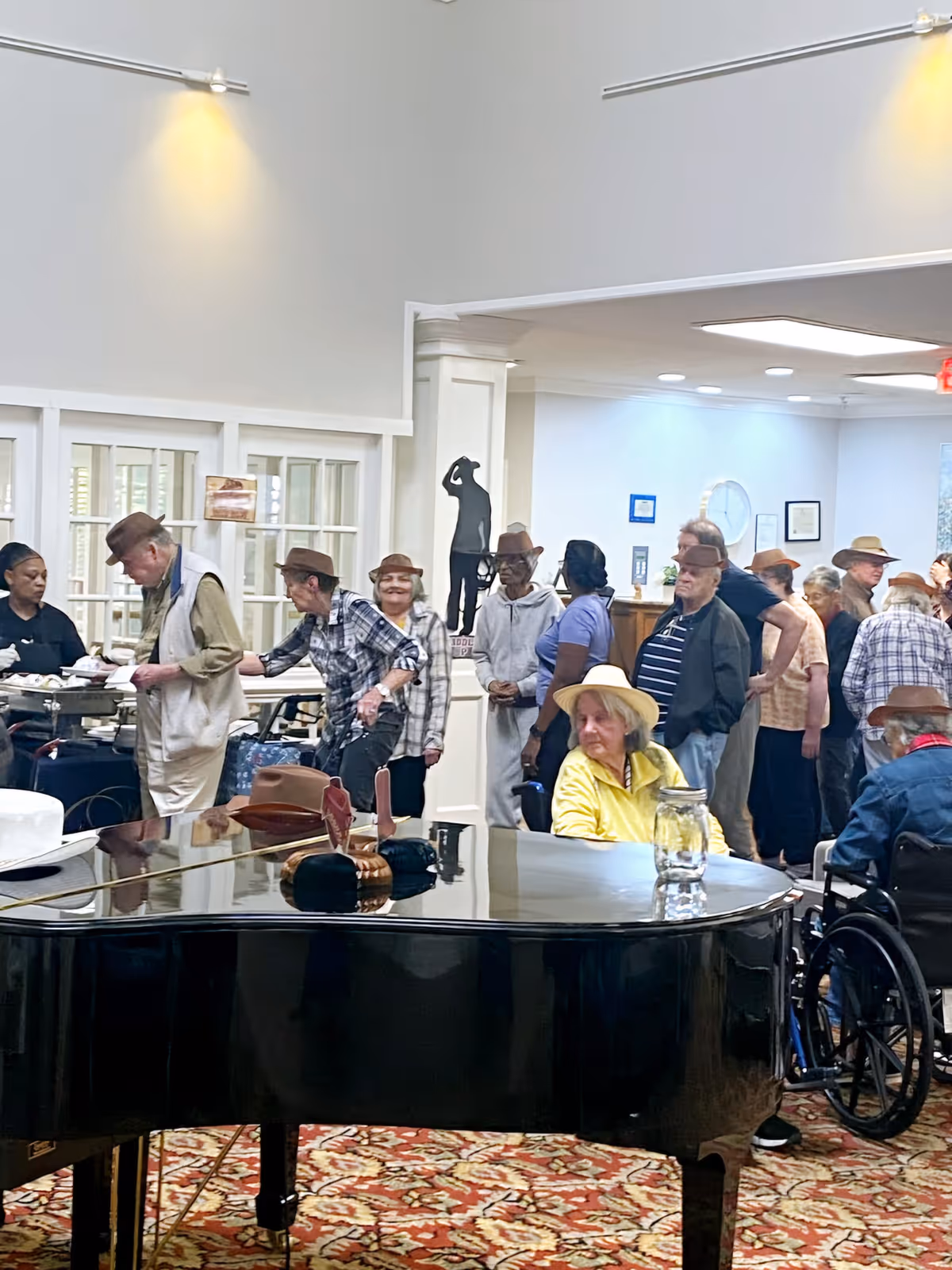 A group of elderly people gathered indoors near a grand piano with a glass of water on it. Some individuals are standing in line, while others are seated, including one person in a wheelchair. The room has white walls, a patterned carpet, and a sculpture in the background.