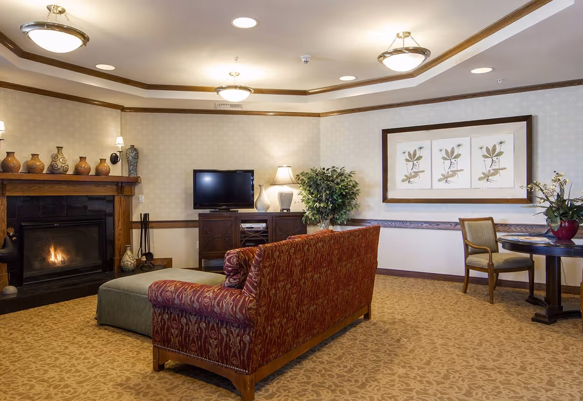 A cozy living room with a patterned red sofa facing a flat-screen TV on a wooden cabinet. To the left, there is a fireplace with decorative vases on the mantel. The room has beige patterned carpet and wallpaper, a framed botanical artwork on the wall, a round table with a chair, and a potted plant near the corner. Ceiling lights illuminate the space warmly.