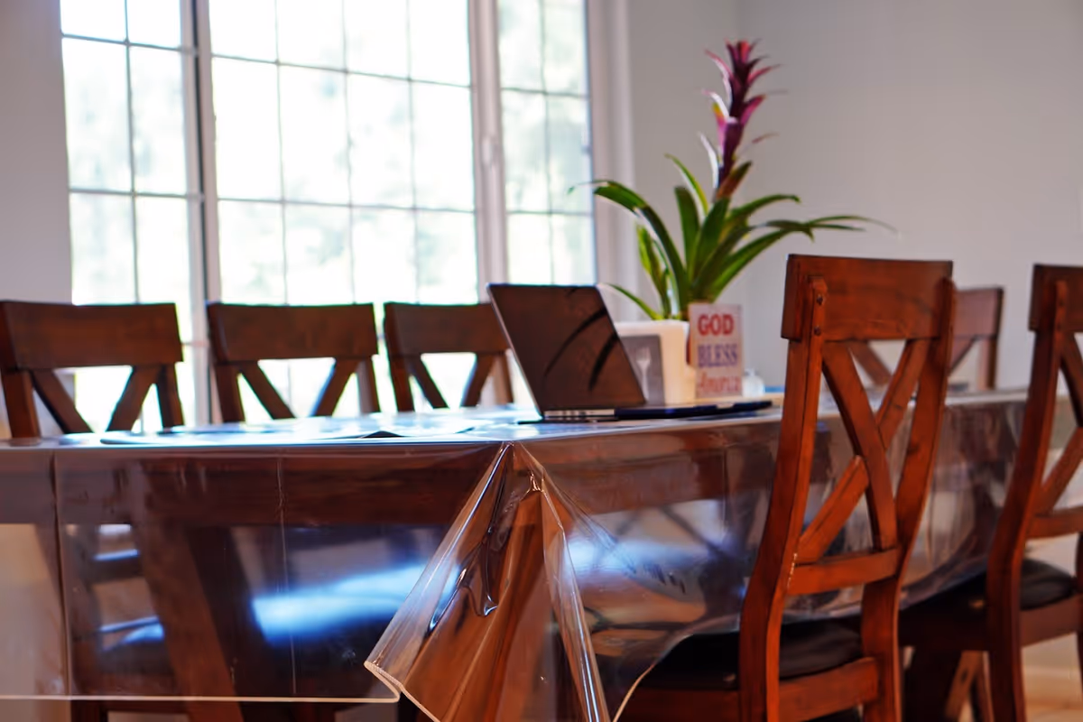A dining table covered with a clear plastic tablecloth surrounded by wooden chairs. On the table, there is a laptop, a potted plant with purple flowers, and a small sign that reads 'GOD BLESS'. Large windows in the background let in natural light.