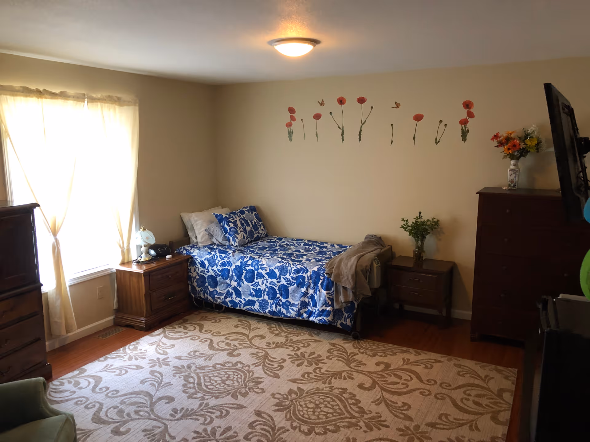 A tidy bedroom with a single bed covered in blue floral bedding, wooden nightstands and dressers, a patterned area rug, and flower wall decals on the wall.