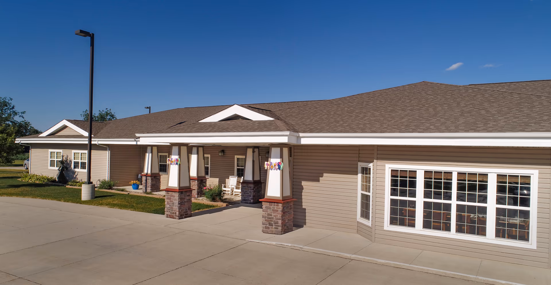 Single-story beige assisted living building with a covered entrance portico, columns, and large windows under a clear blue sky.