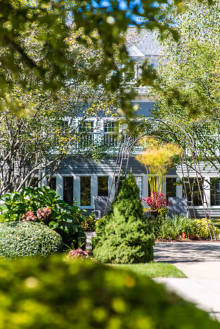 A lush garden area with various green shrubs, trees, and flowering plants in front of a building with multiple windows. There is a metal archway structure in the garden and a paved walkway leading through the greenery.
