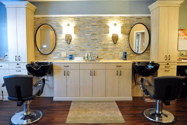 Interior view of a salon area with two black salon chairs in front of black wash basins. The background features a beige cabinet with multiple drawers and cupboards, a small sink in the center, two oval mirrors mounted on a stone-tiled wall, and two wall sconces providing light above the sink area. The floor is wooden with a small patterned rug in front of the cabinet.