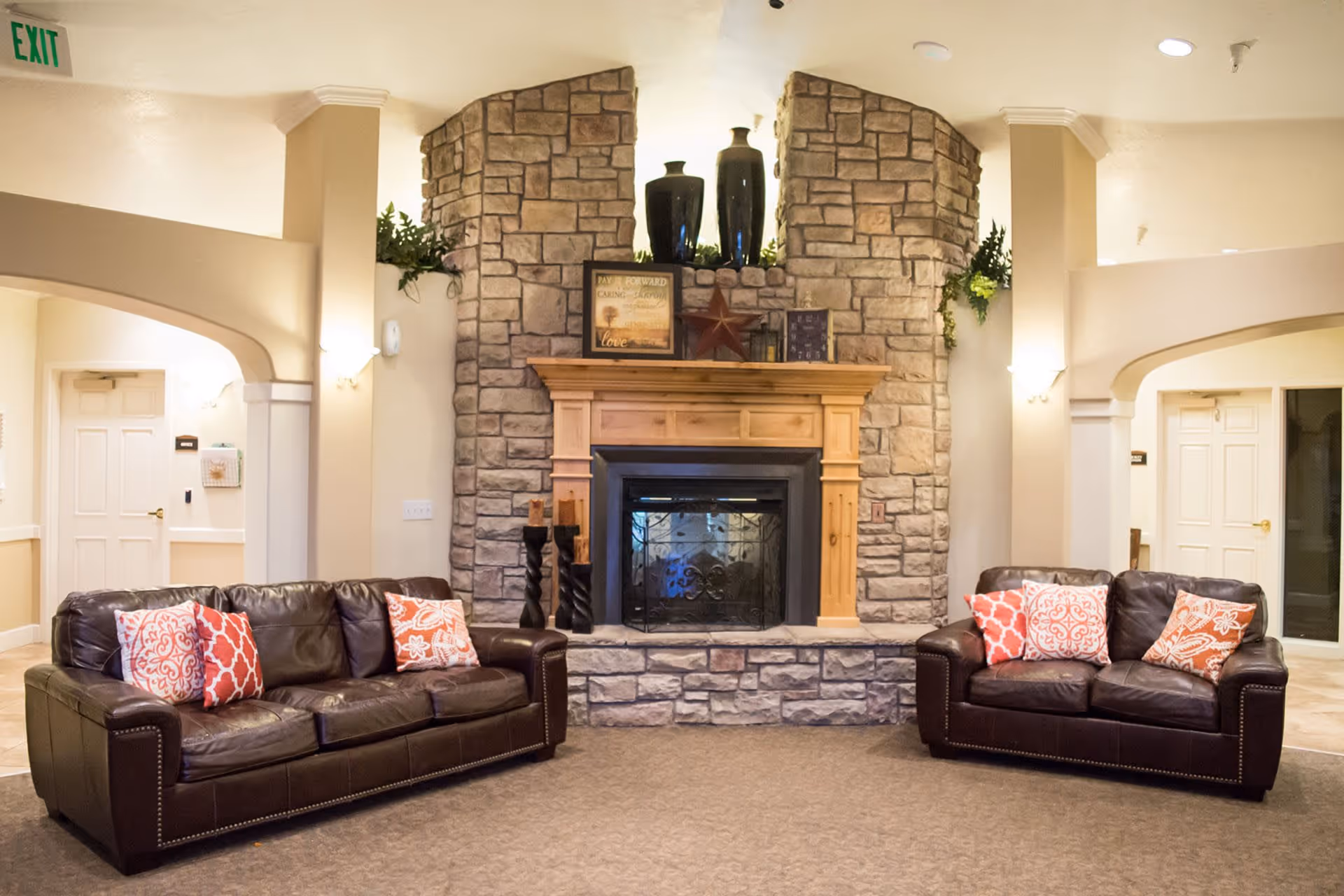A cozy living room area in an assisted living facility featuring two dark brown leather sofas with red and white patterned throw pillows facing a stone fireplace with a wooden mantel. The mantel is decorated with vases, framed pictures, and a star ornament. The room has beige walls, carpeted floor, and soft lighting with wall sconces. There are archways and doors visible in the background.