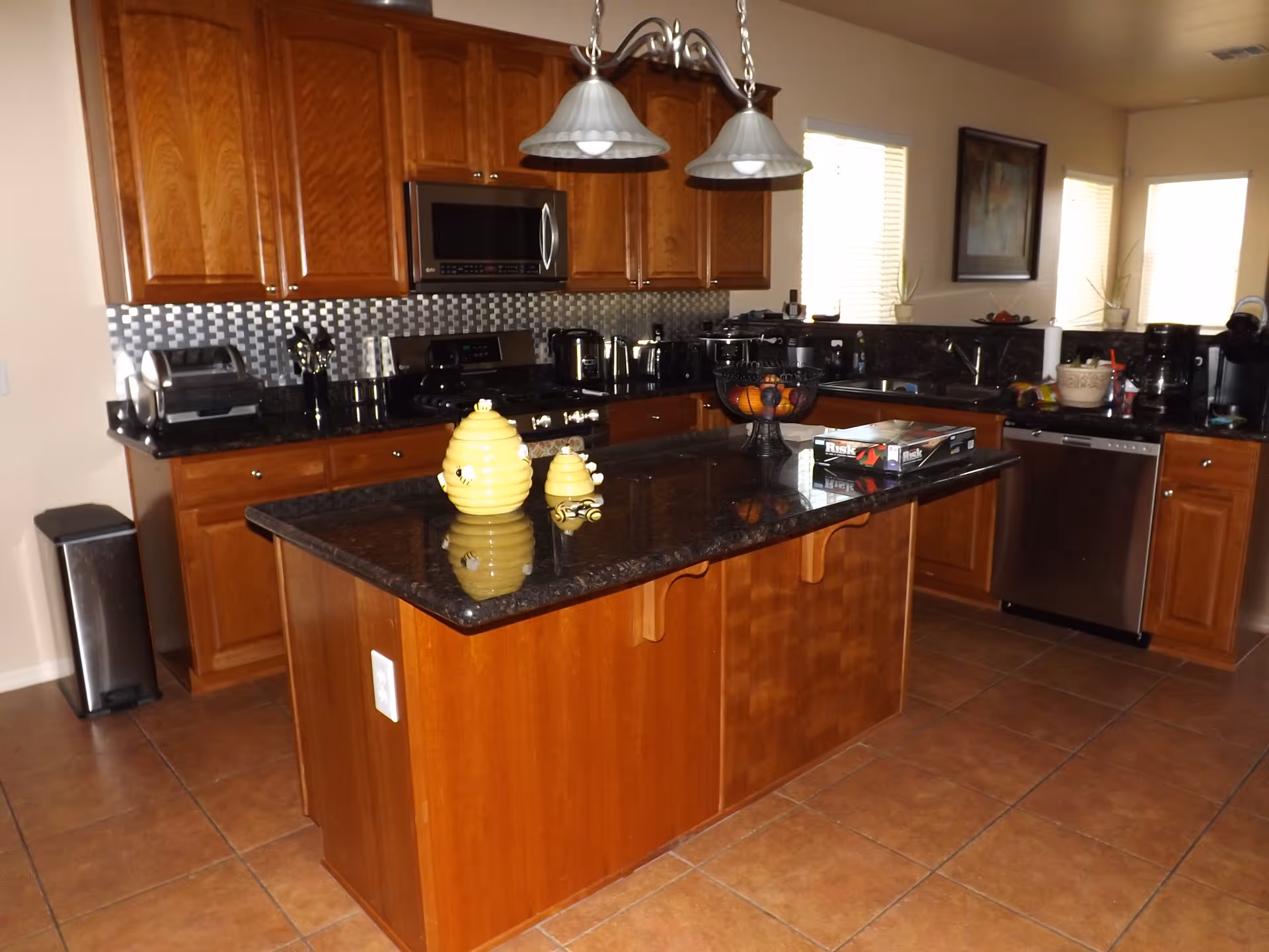 A kitchen with wooden cabinets, a black granite countertop island with decorative yellow bee-themed containers, and stainless steel appliances including a microwave, stove, and dishwasher. The kitchen has tiled flooring, a trash bin, and two hanging pendant lights above the island. There are windows with blinds and a framed picture on the wall.