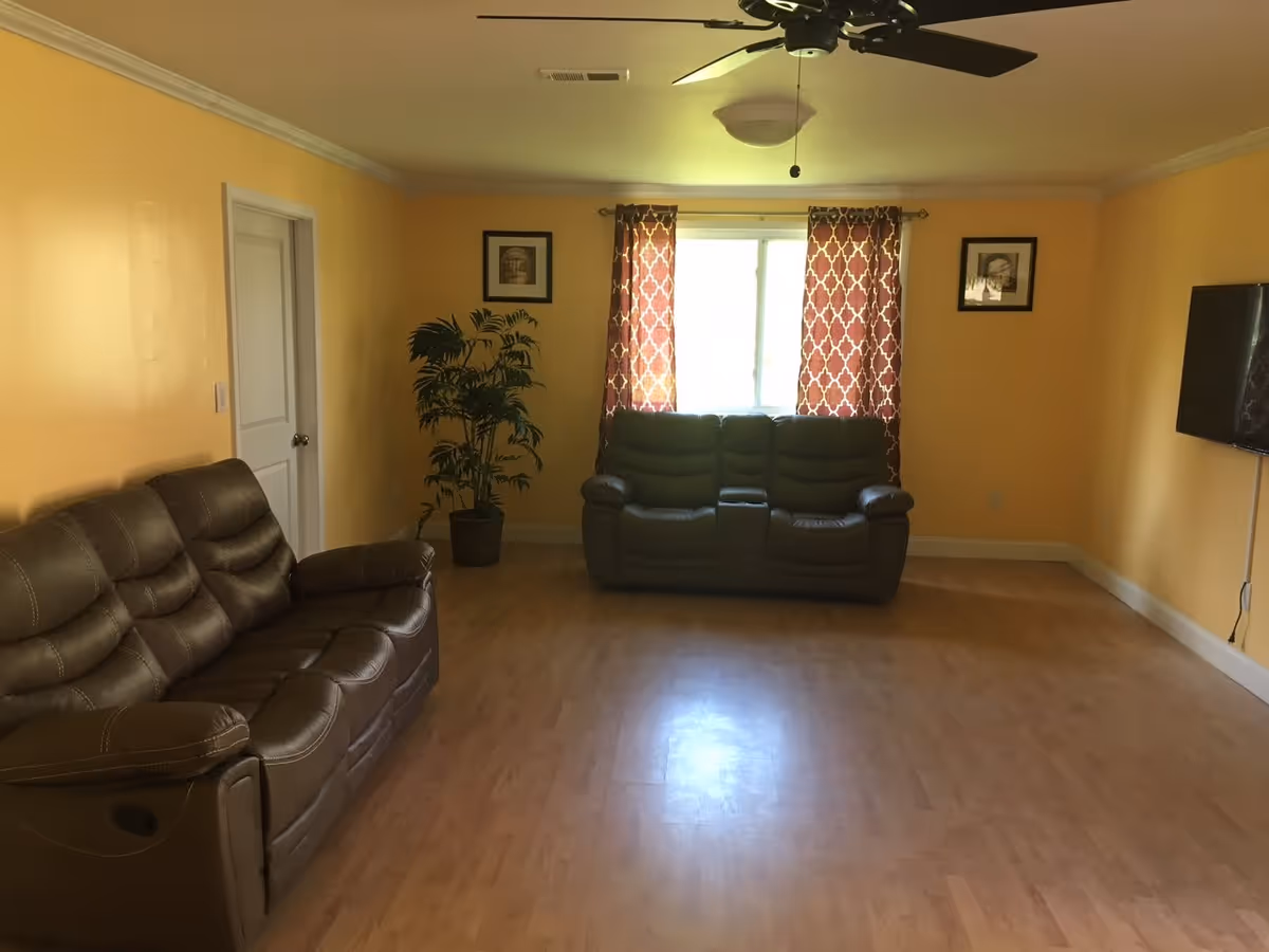 A living room with yellow walls and light wood flooring featuring two brown leather sofas, a potted plant near a white door, a window with red patterned curtains, two framed pictures on the wall, a ceiling fan, and a wall-mounted flat-screen TV.